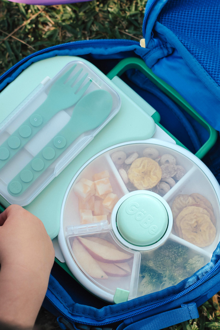 Green fork and spoon in their case tucked into a lunchbox beside a snack spinner