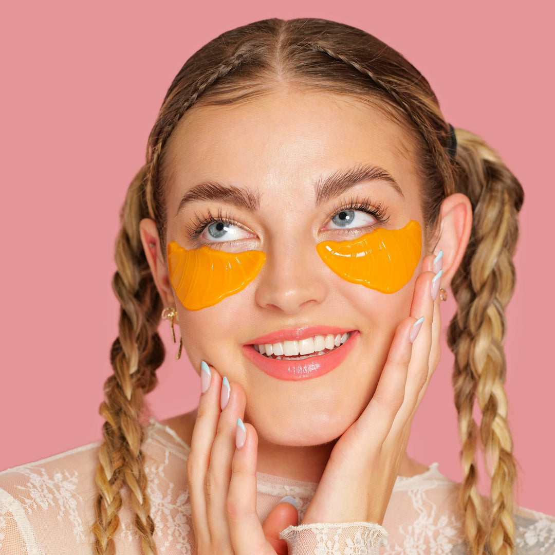 Model glancing up while wearing shiny croissant eye patches, playful depuffing for a girls spa day