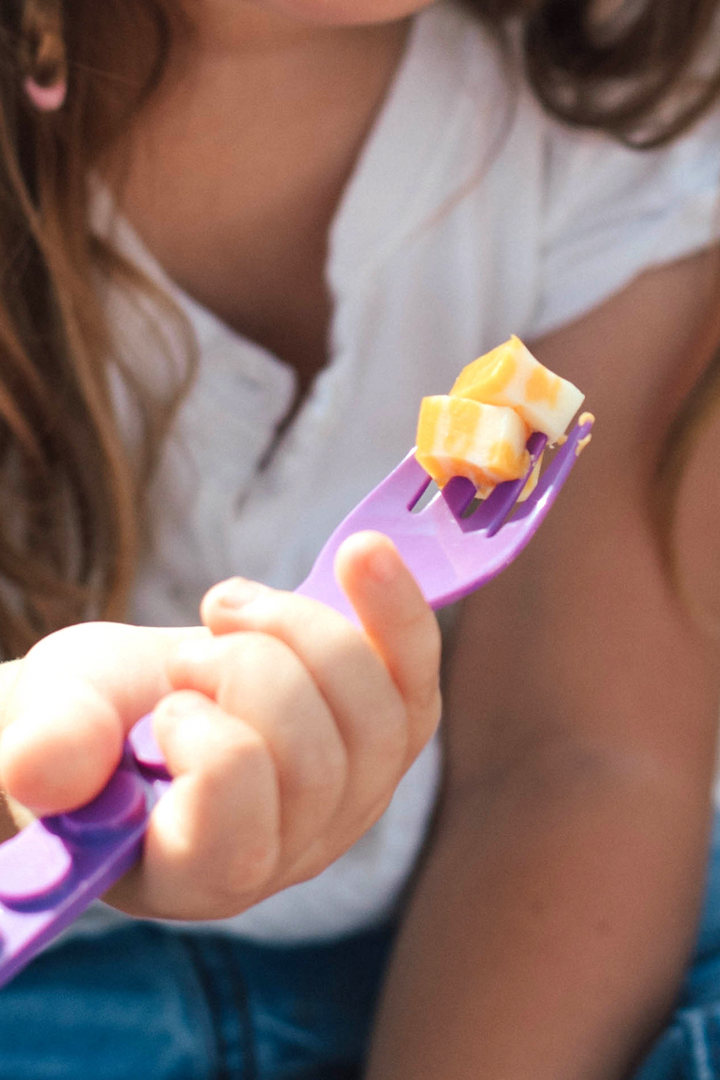Child's hand holding a purple GoBe fork with a piece of fruit on it