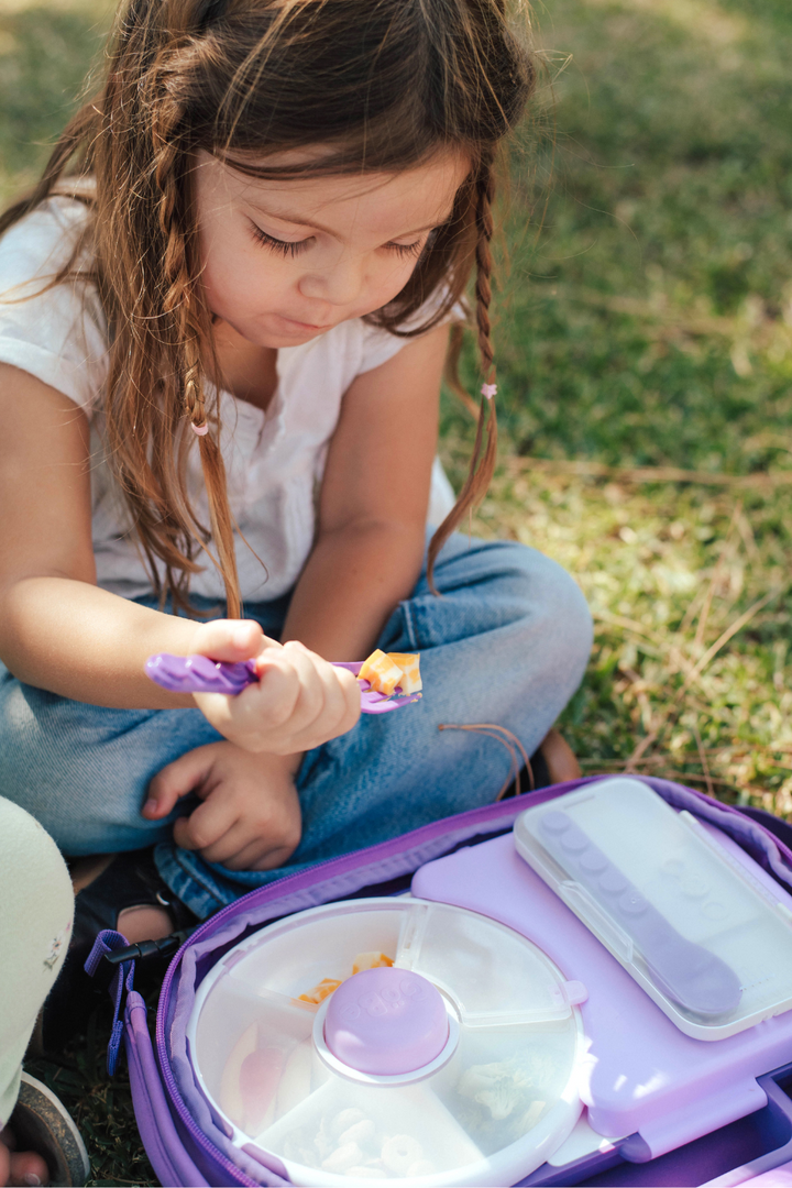 Girl on the grass clicking utensils together beside an open purple lunch bag