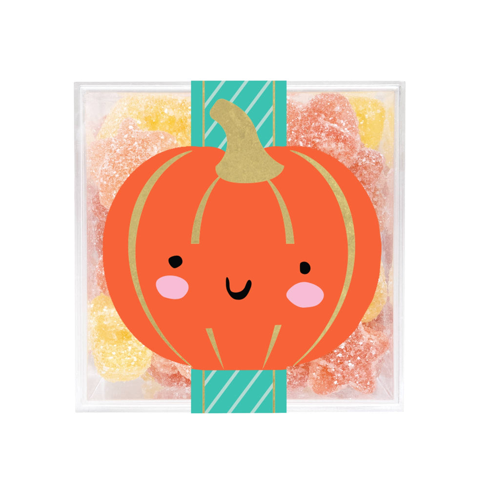 A clear square gift box filled with assorted sugar-coated gummy candies, featuring a large orange pumpkin illustration with a smiling face and pink cheeks wrapped around the front.
