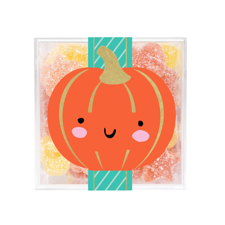 A clear square gift box filled with assorted sugar-coated gummy candies, featuring a large orange pumpkin illustration with a smiling face and pink cheeks wrapped around the front.