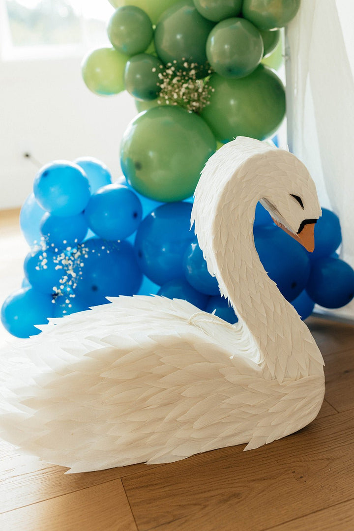 White swan-shaped pinata on a wooden floor with green and blue balloon arches in the background.