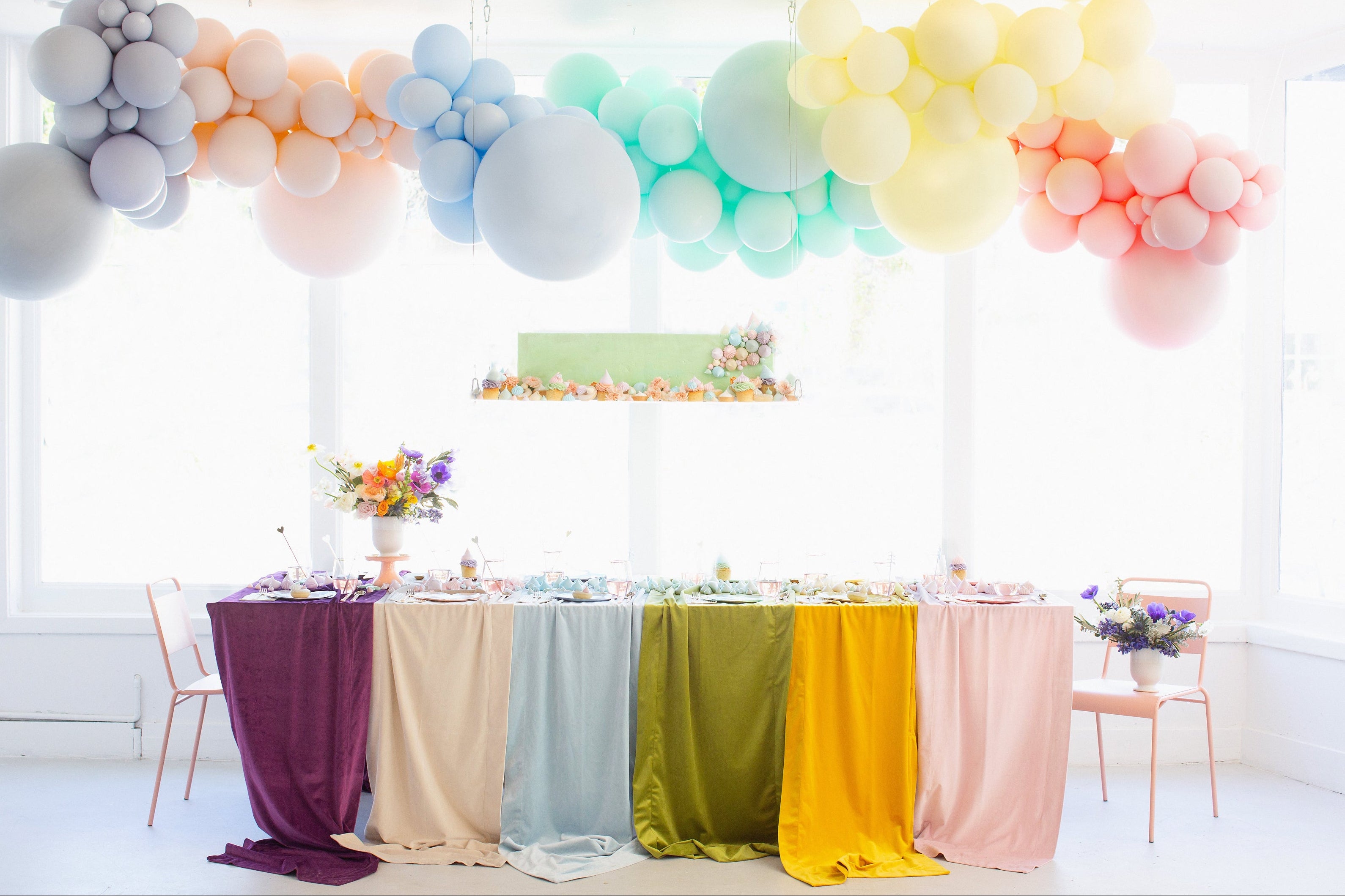 Pastel rainbow balloon garland over a party table with jewel-tone velvet linens and a cupcake display for a birthday celebration