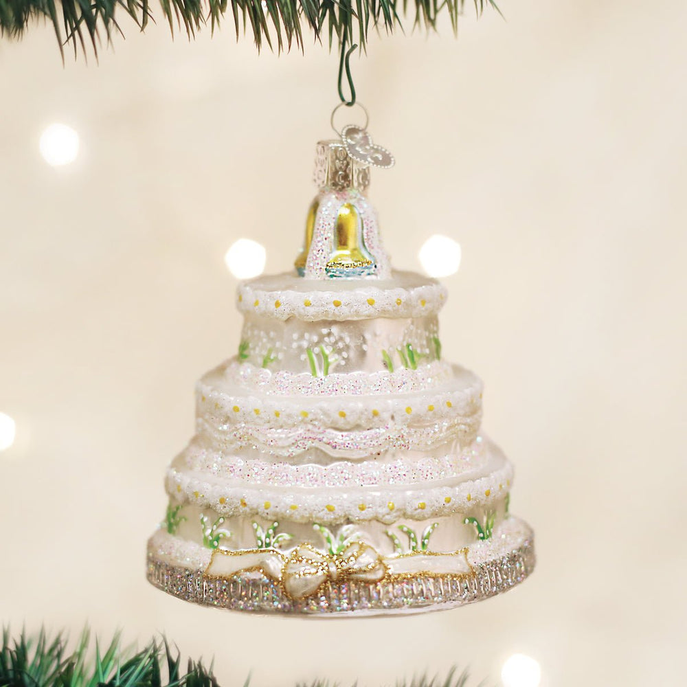 Three-tier white wedding cake glass ornament with gold bells on top, hanging on a Christmas tree