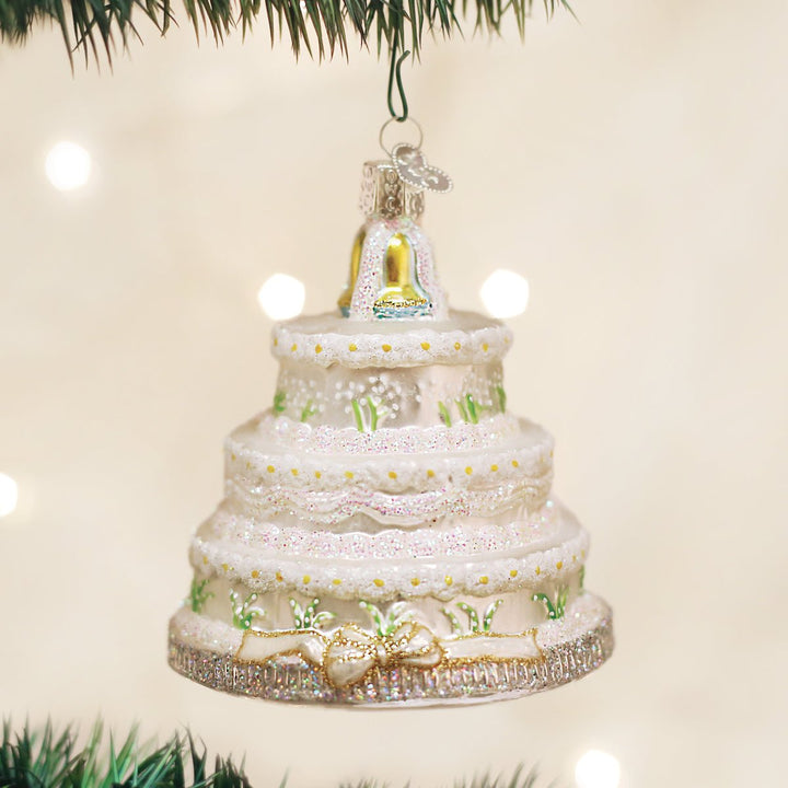 Three-tier white wedding cake glass ornament with gold bells on top, hanging on a Christmas tree