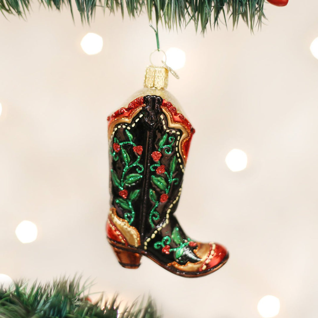 Holly berry cowboy boot ornament hanging on a lit Christmas tree with soft bokeh lights