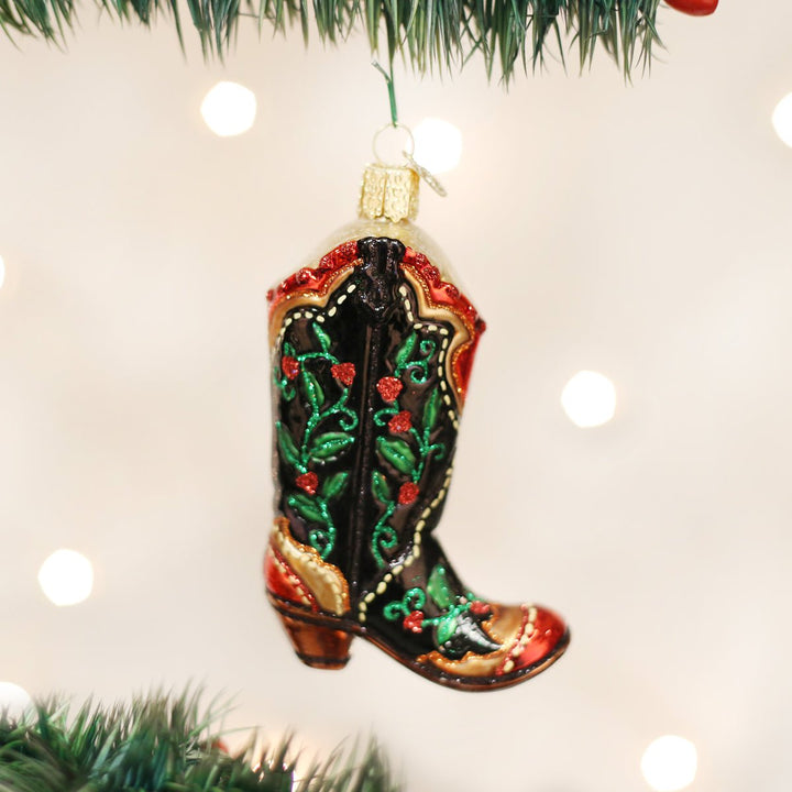 Holly berry cowboy boot ornament hanging on a lit Christmas tree with soft bokeh lights