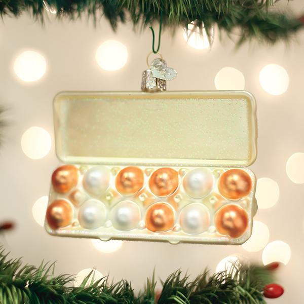 Open egg carton glass ornament filled with brown and white eggs, hanging on a lit Christmas tree
