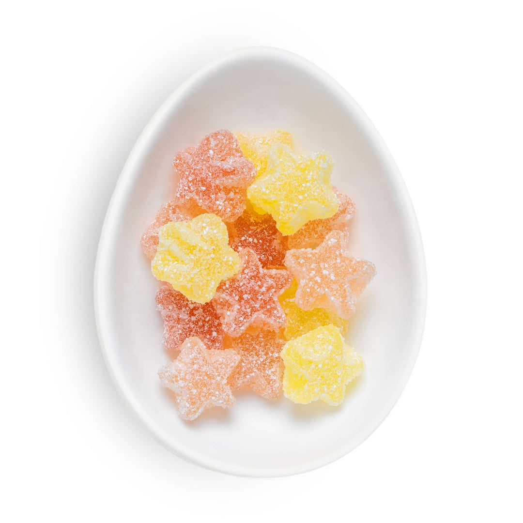 Star-shaped sugar-dusted gummy candies in yellow and pink, arranged in a white ceramic egg-shaped dish on a white background.