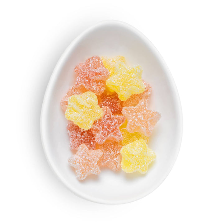 Star-shaped sugar-dusted gummy candies in yellow and pink, arranged in a white ceramic egg-shaped dish on a white background.