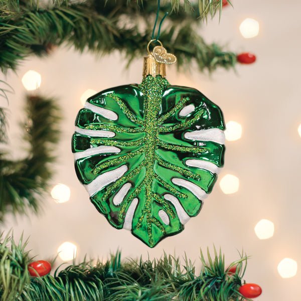 Green monstera leaf ornament hanging on a lit Christmas tree with warm bokeh lights and red berries