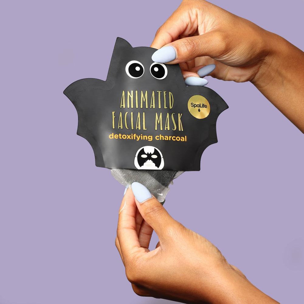 Hands pulling the charcoal sheet mask from its black bat pouch on a lavender background for Halloween skincare