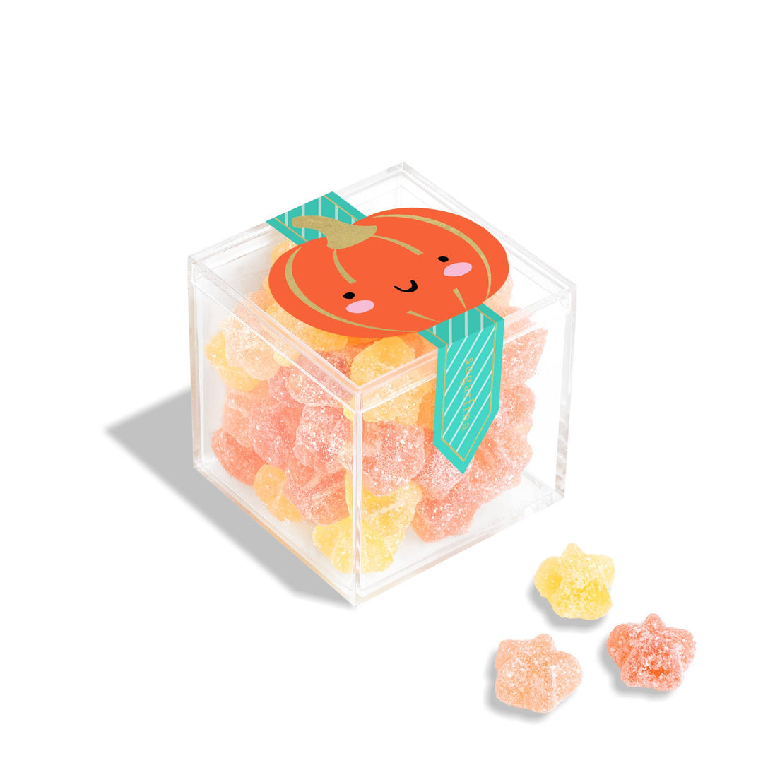 A clear acrylic cube container filled with colorful gummy candies, topped with a smiling orange pumpkin label and teal ribbon, on a white background with three loose star-shaped gummies in front.