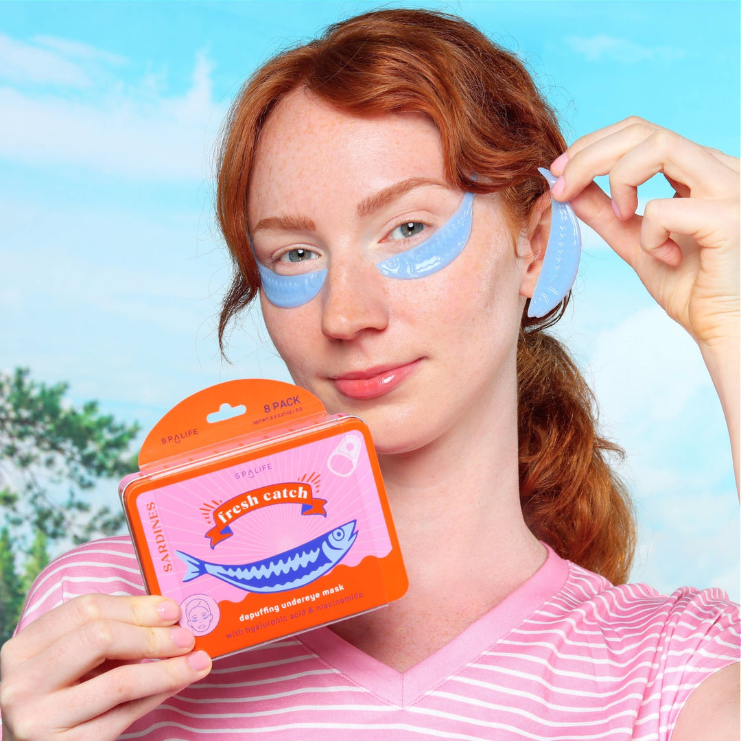 Redheaded model applying blue sardine under eye patches against a lakeside backdrop, a playful summer spa moment