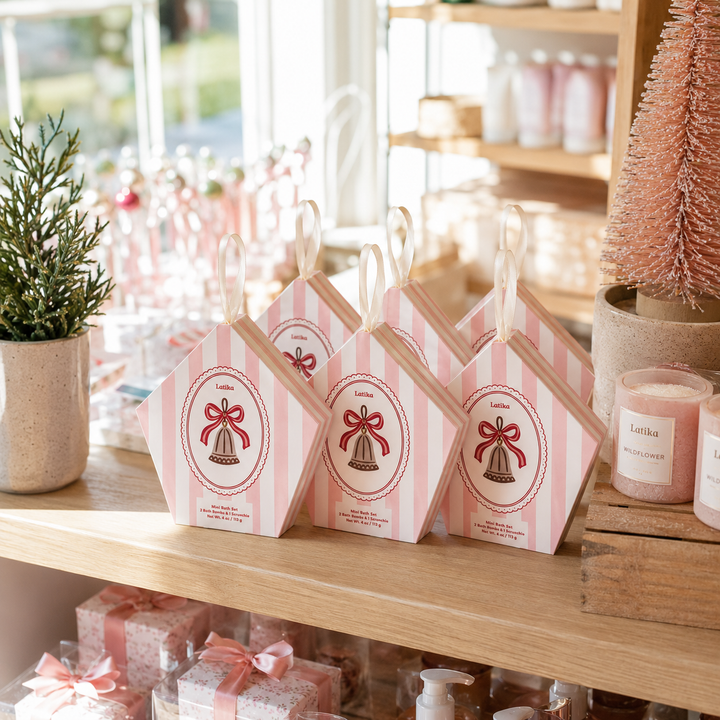 Three pink striped bell ornament boxes on a boutique shelf with candles and a mini tree for holiday shopping