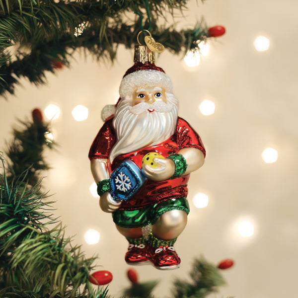 Santa pickleball glass ornament in a red tee and green shorts hanging on a lit Christmas tree