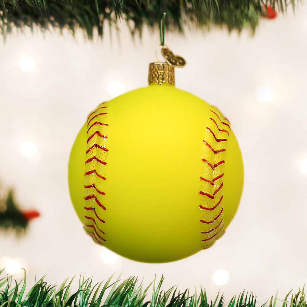 Bright yellow softball ornament hanging from a green tree branch with soft lights glowing behind