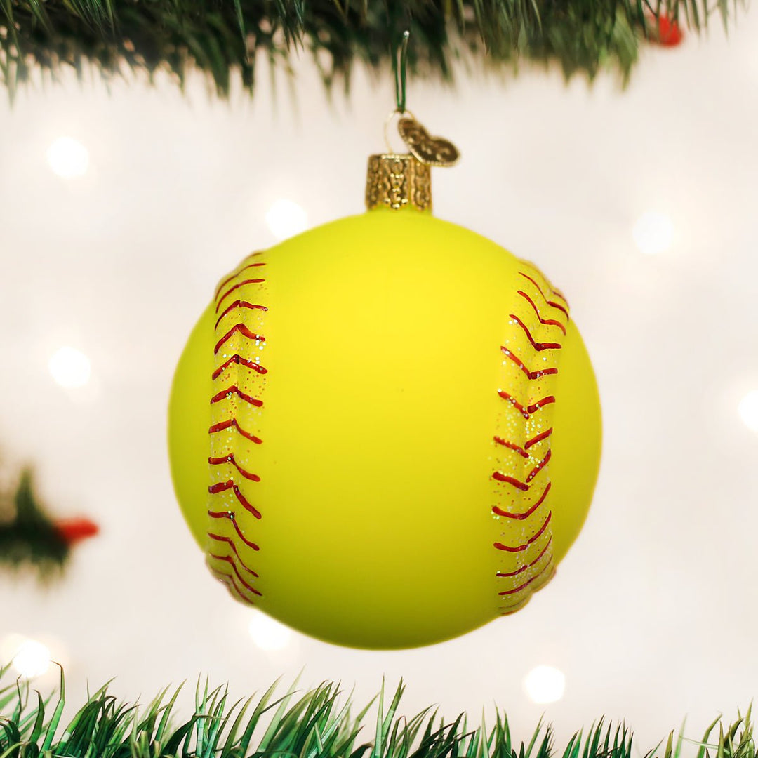 Bright yellow softball ornament hanging from a green tree branch with soft lights glowing behind