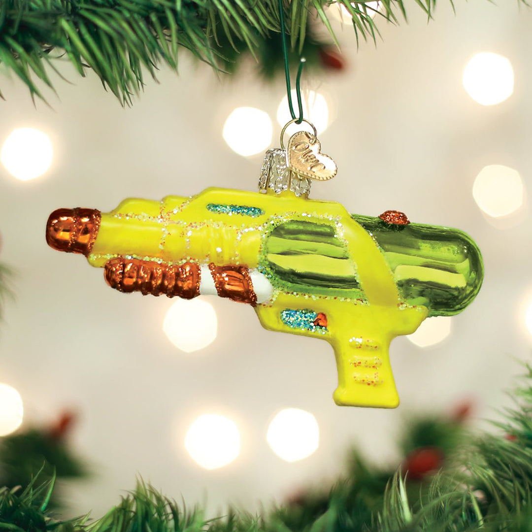 Yellow and green squirt gun ornament hanging on a lit Christmas tree with warm bokeh lights