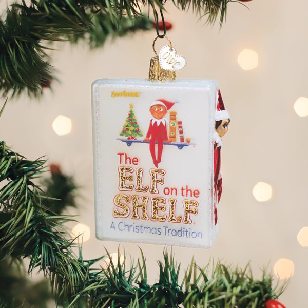 The Elf on the Shelf book ornament hanging on a Christmas tree with warm lights behind it