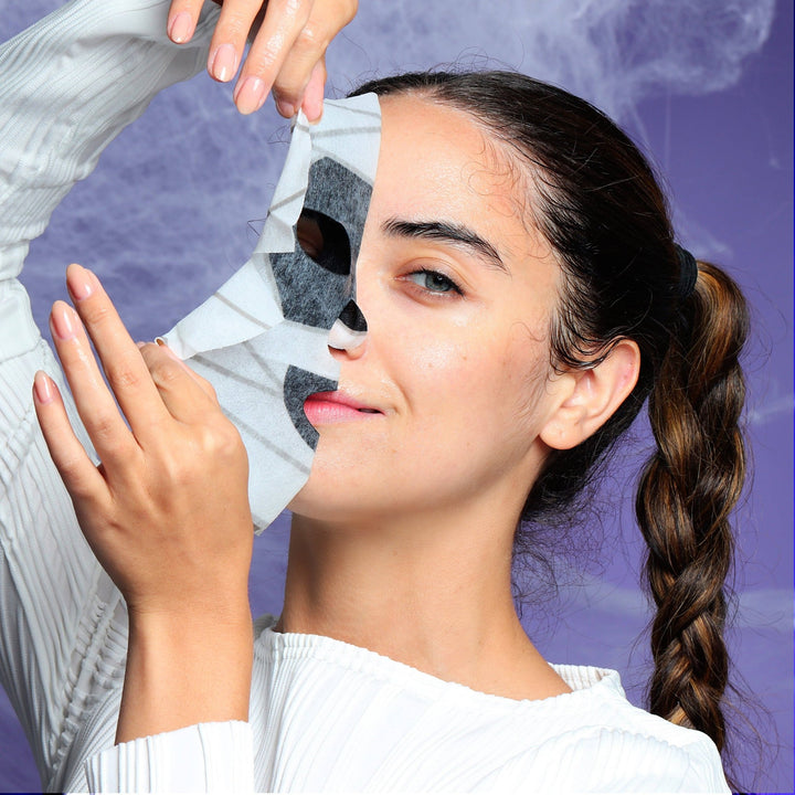 Model lifting the mummy sheet mask off half her face against a cobweb backdrop during a Halloween spa night