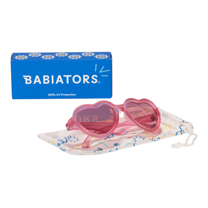 Fuchsia heart kids sunglasses folded on a printed storage pouch beside the Babiators box.
