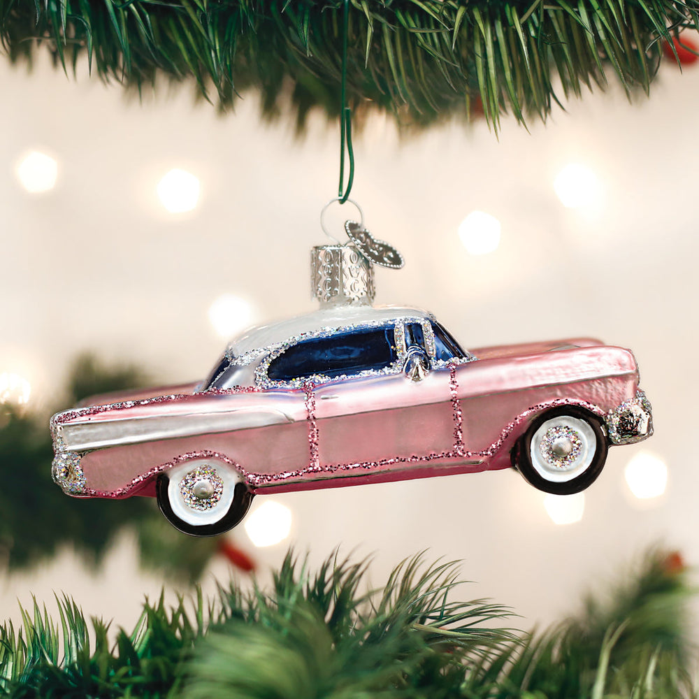 Pink classic car Christmas ornament hanging from a pine branch against glowing holiday lights