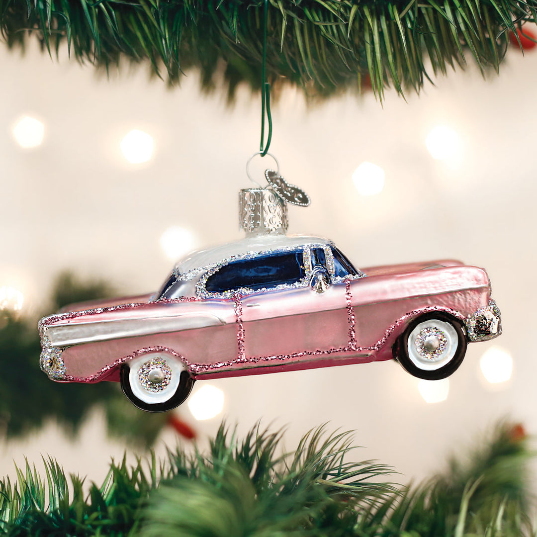 Pink classic car Christmas ornament hanging from a pine branch against glowing holiday lights