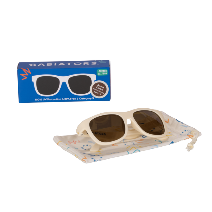 Cream aviator kids sunglasses folded on a printed storage pouch beside the Babiators Originals box.