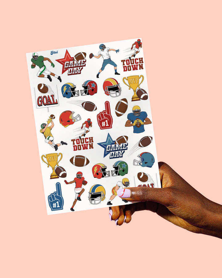 A hand holding one 8 by 6 inch sheet of football temporary tattoos with helmets and footballs