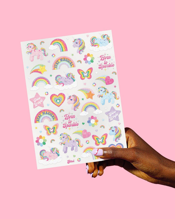 Hand holding one sheet of pastel glitter unicorn tattoos with rainbows, hearts, stars, and Shine Bright slogans