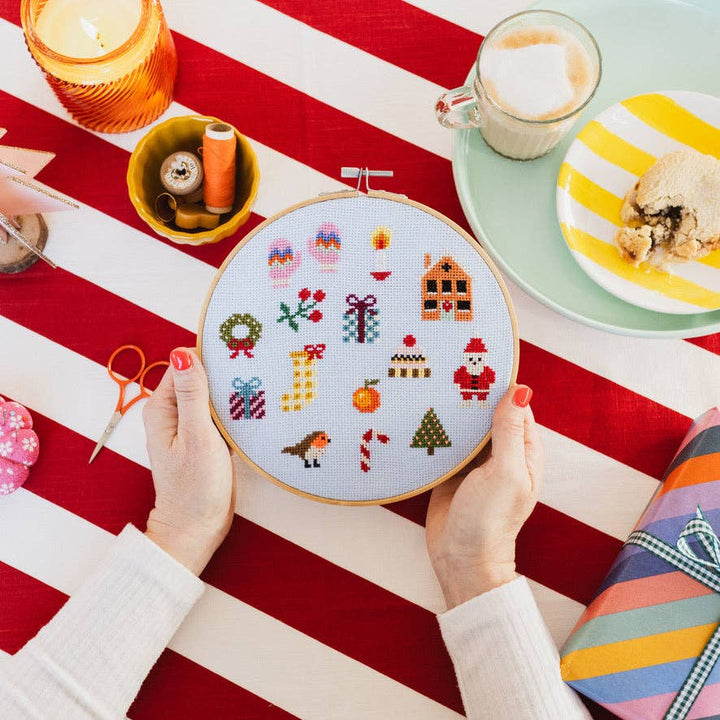 Hands holding colorful Christmas cross stitch sampler over a red striped tablecloth