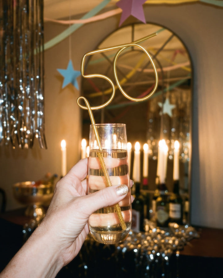 Hand holding a champagne flute with the gold 50 straw beside lit candles