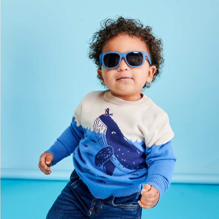 A curly-haired toddler in a whale sweater wearing blue aviator sunglasses against a light blue backdrop.