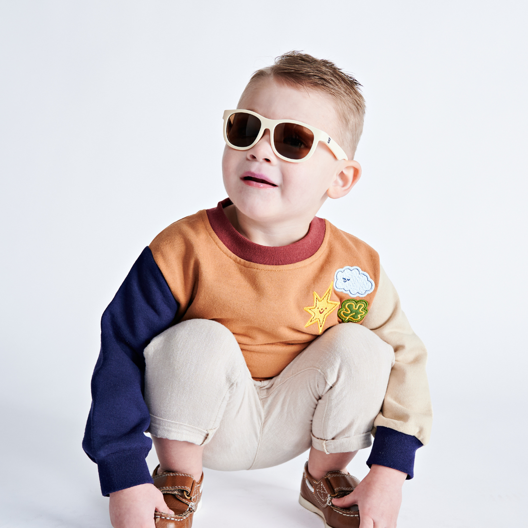 A young boy in a colorblock sweater crouching and wearing cream aviator sunglasses against a white backdrop.