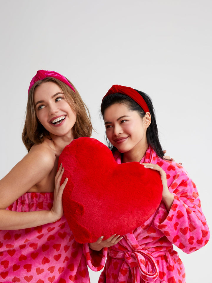 Two women in pink heart-patterned robes and matching headbands hugging a large red heart-shaped plush pillow against a light gray studio background.