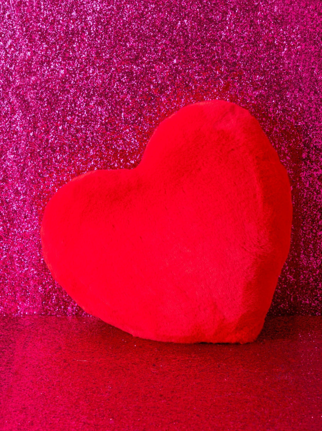 A bright red fuzzy heart-shaped cushion or pillow standing against a sparkling hot pink glitter background.