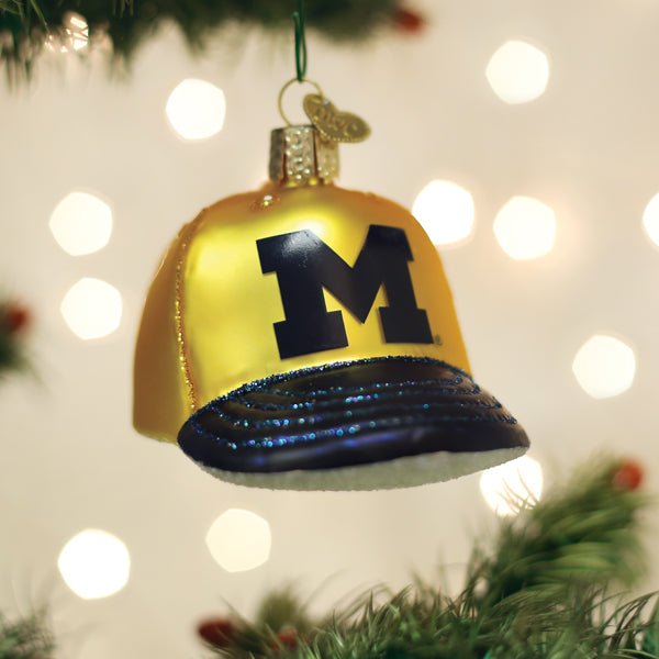 Maize and navy Michigan Wolverines cap ornament hanging on a Christmas tree with warm twinkle lights