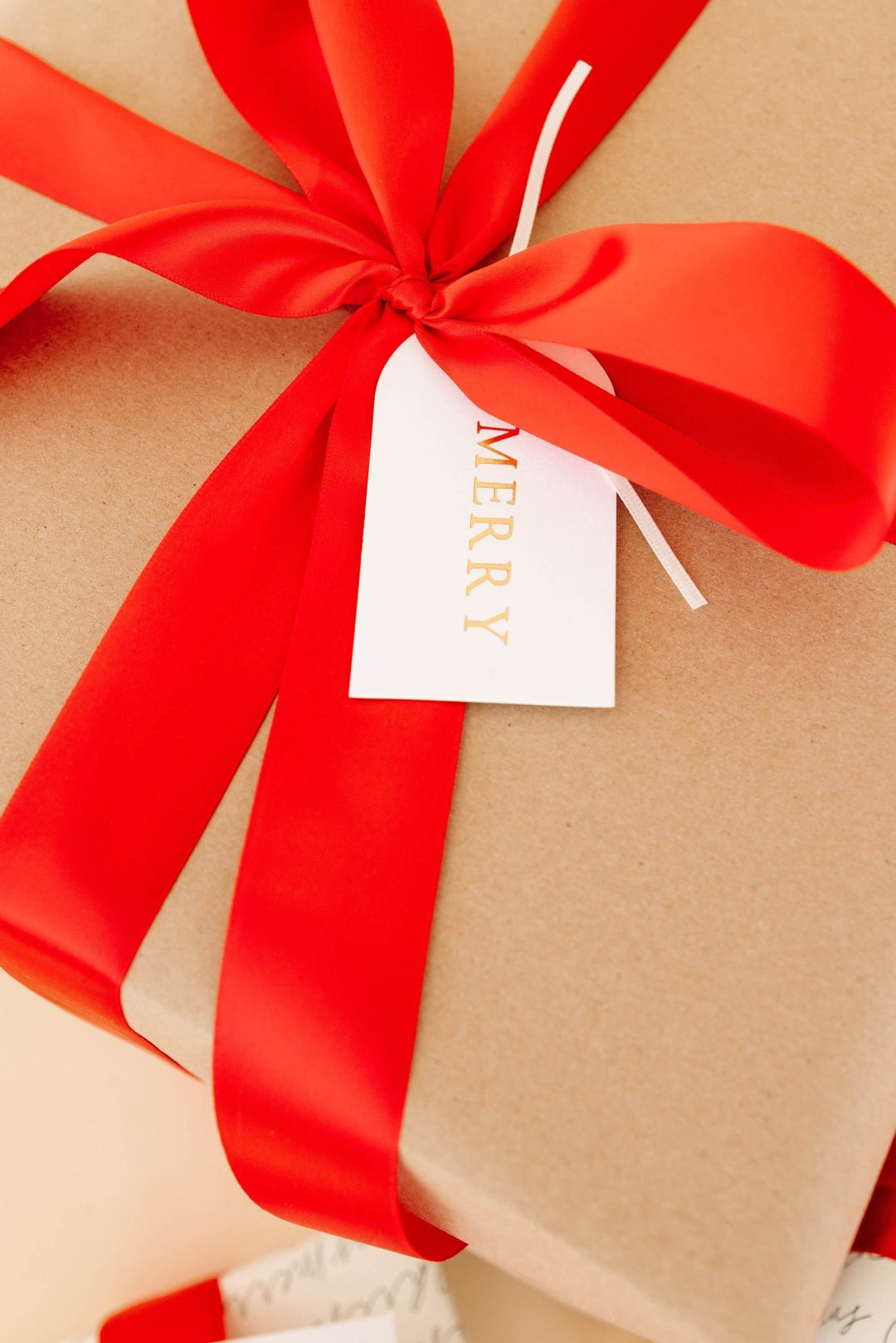 Arch MERRY tag in gold foil on a kraft paper gift tied with a bright red satin bow
