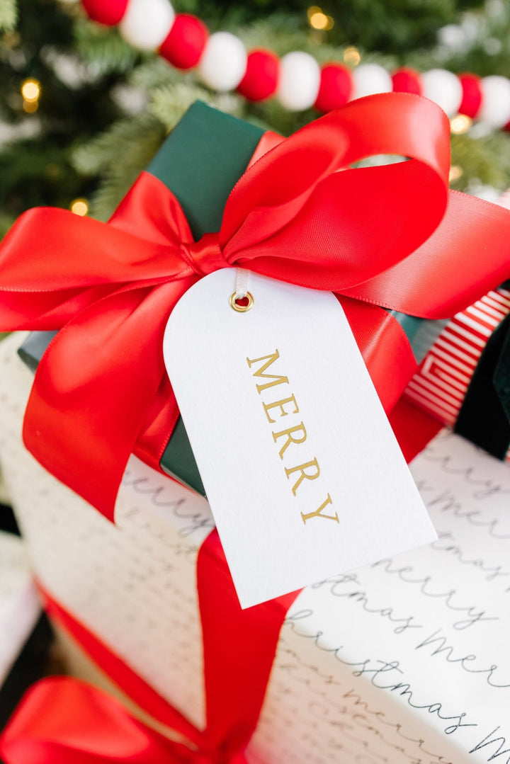 Arch Merry gift tag on a red satin bow by a Christmas tree with a pom pom garland behind