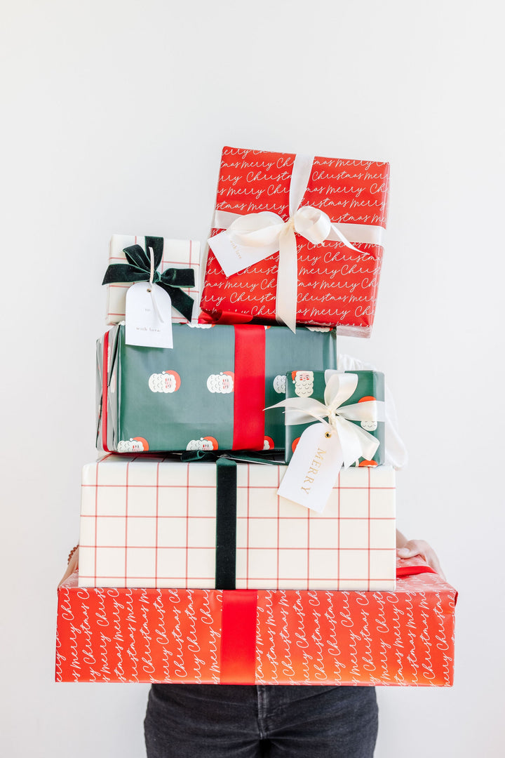 Arms holding a tall stack of Santa print, grid, and red script wrapped gifts against a white wall