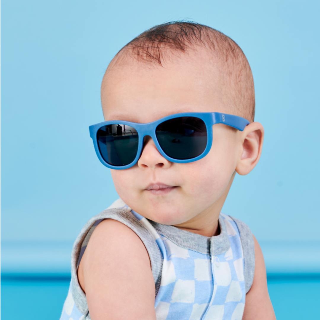 A baby in a checkered tank top wearing blue aviator sunglasses against a light blue backdrop.