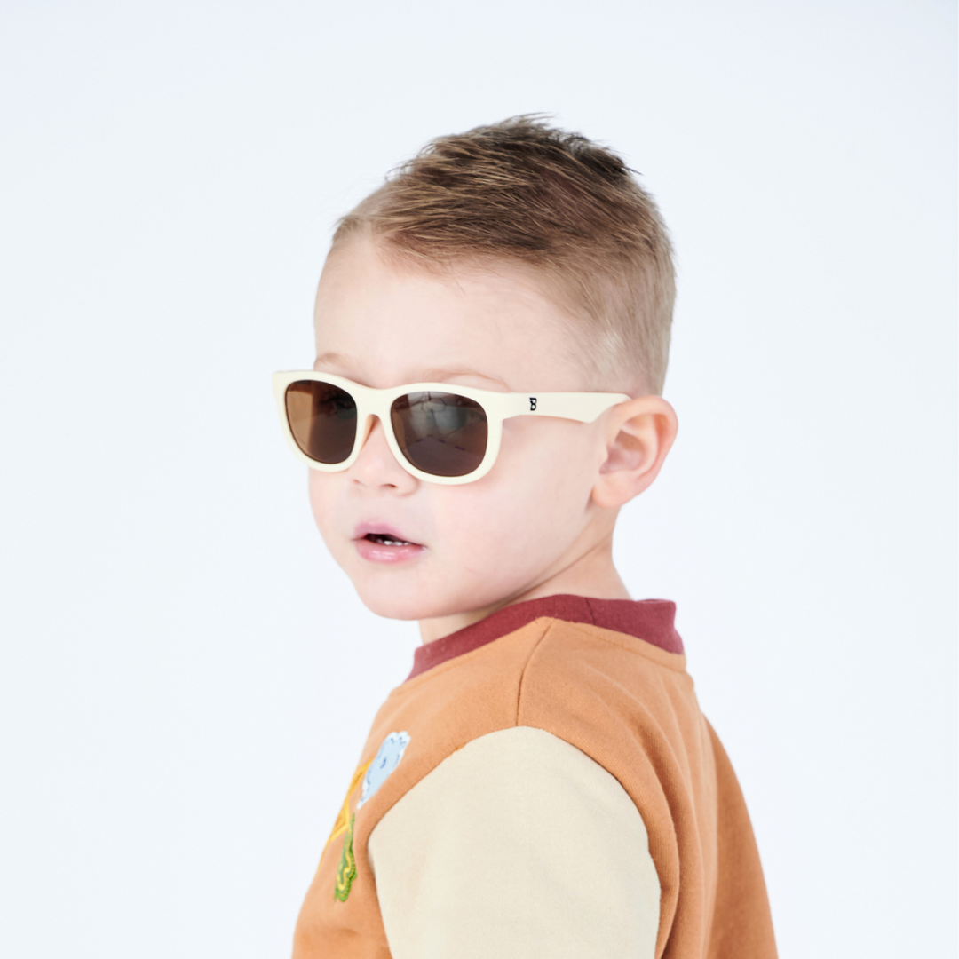 Side profile of a young boy wearing cream aviator sunglasses against a white backdrop.