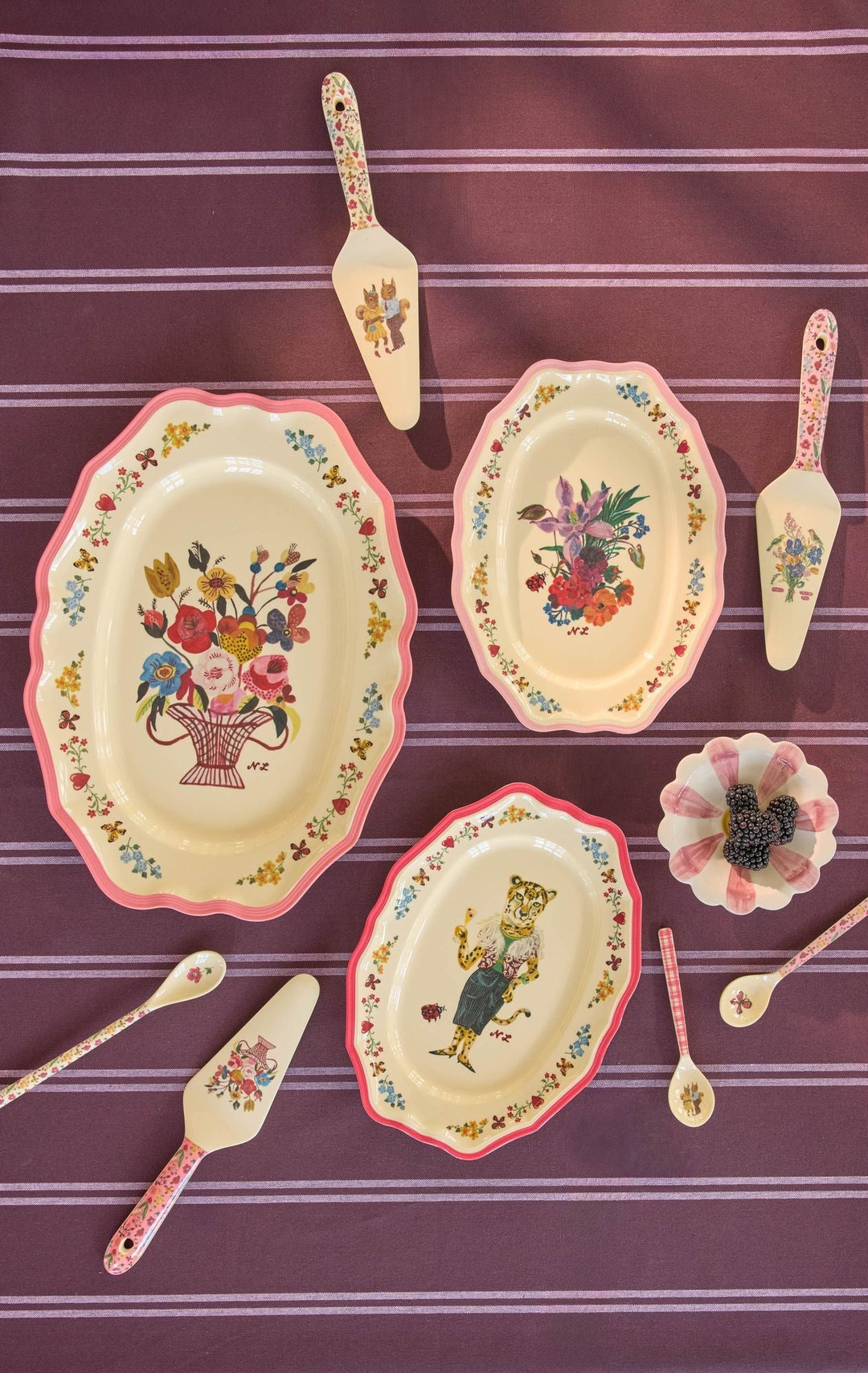 Nathalie Lété serving dishes and matching floral cake servers laid out on a plum striped tablecloth