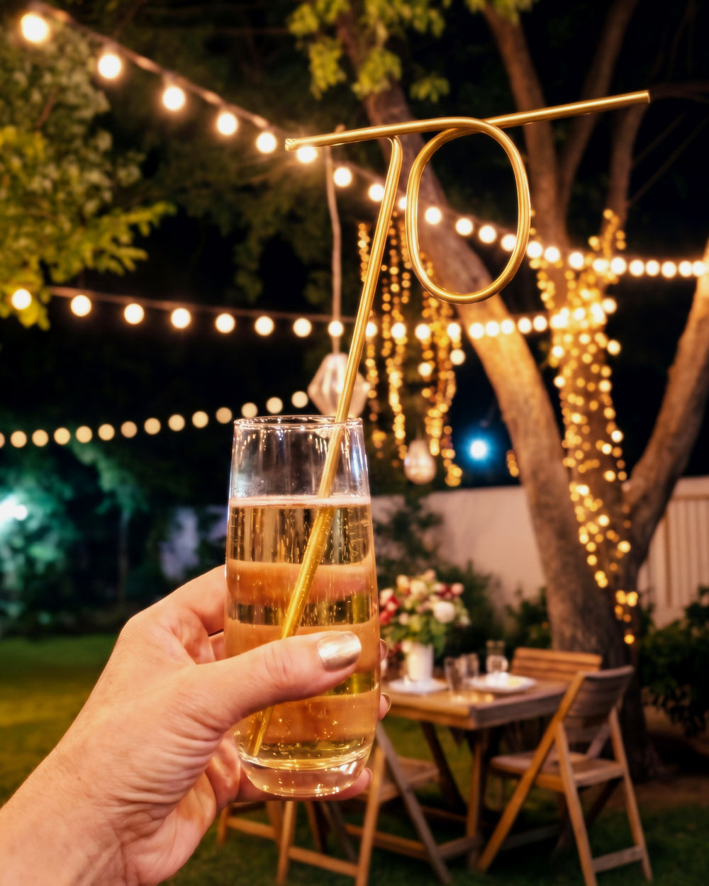 Hand holding a champagne flute with the gold 70 straw at an outdoor party