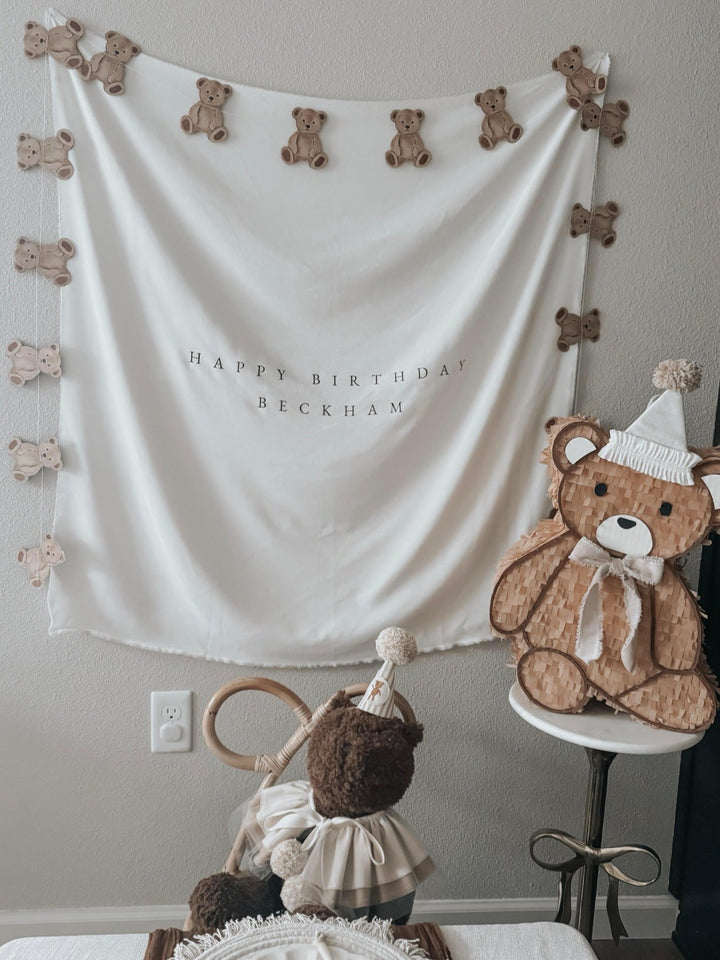Teddy bear piñata on a stand beside a bear garland and backdrop at a neutral teddy bear first birthday party