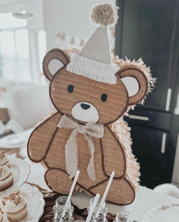 Fringed teddy bear piñata in a party hat with a ribbon bow, styled on a teddy bear birthday dessert table with cupcakes