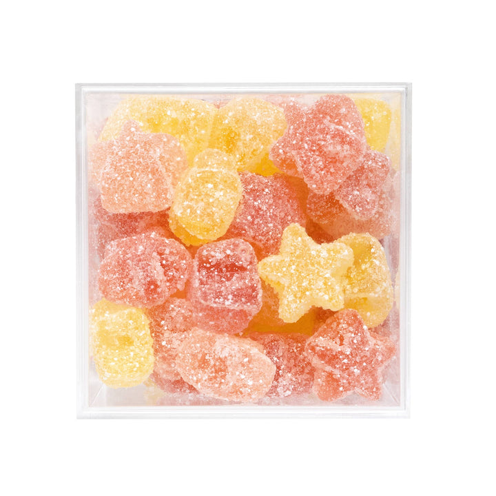 A clear square plastic container filled with sugar-coated gummy candies in yellow and pink-orange tones, photographed on a plain white background.