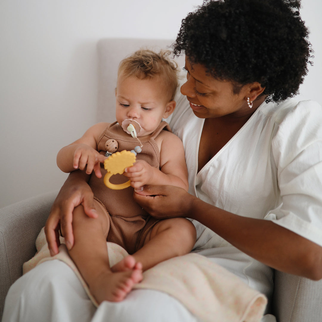 Baby on a parent's lap holding a yellow Mushie lion ring teether